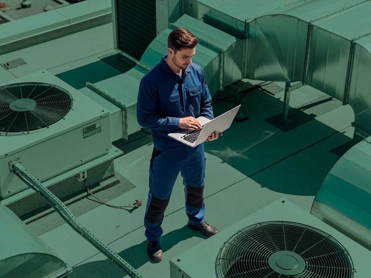 Servicetechniker mit Laptop auf einem Gebäudedach zwischen Lüftungs- und Klimaanlagen