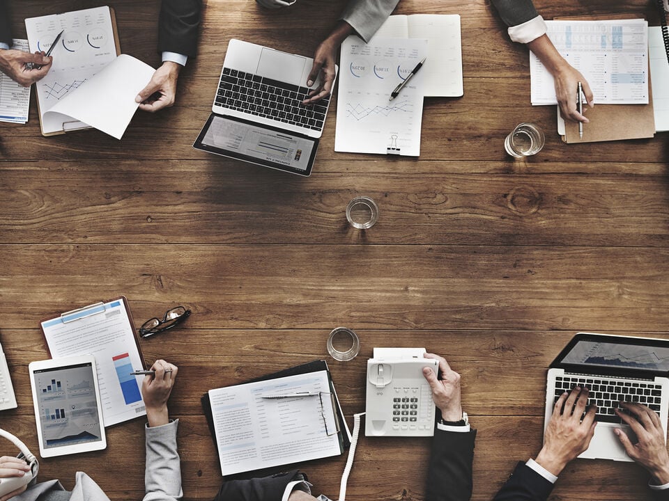 Top-down view of people working together with laptops, tablets, and documents on a desk.