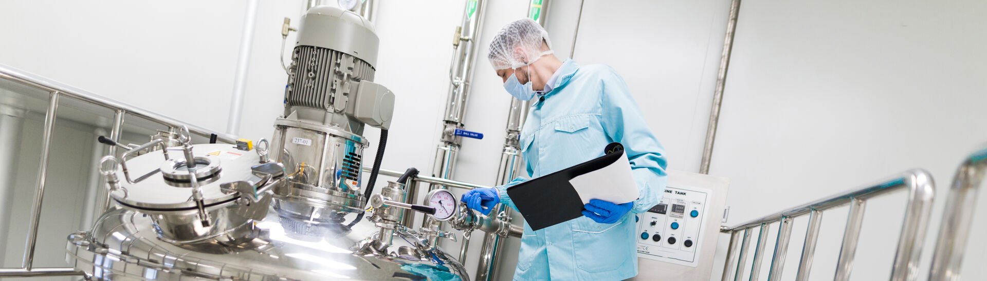 Widescreen picture, scientist in blue lab suit and mask, examining manufacturing equipment.