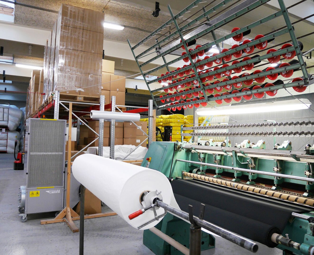 A manufacturing area featuring a large machine, rolls of material and stacked boxes in the background.